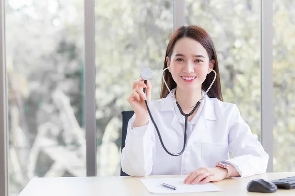 専門のアジア人女性医師は 健康診断のコンセプトで病院でカメラを見ながら オフィスの部屋で医療コートや聴診器を身に着けています — ストック写真