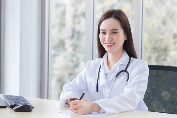 専門のアジア人女性医師は 紙に何かを書いている間に医療用のコートと聴診器を着用し 彼女は健康診断の概念で病院でカメラを見てオフィスの部屋で働いています — ストック写真