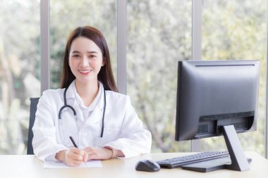 Profesyonel Asyalı kadın doktor kağıda bir şey yazarken ve ofis odasında çalışırken tıbbi ceket ve steteskop giyer ve sağlık kontrolü konseptiyle hastanede kameraya bakar..
