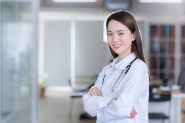 Asyalı profesyonel kadın doktor kolları bağlı duruyor. Hastanedeki muayene odasında beyaz bornoz ve steteskop giyiyordu..