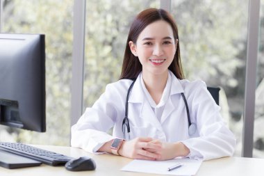 Asyalı profesyonel doktor oturmuş, beyaz bir bornoz ve steteskop takmış gülümsüyor ve hastane sağlık hizmetleri konseptinde bir hastayı bekliyor..