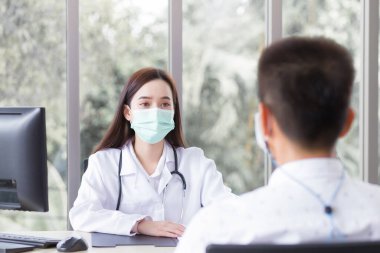 Asyalı profesyonel kadın doktor hastane muayene odasında bir hastayı muayene ederken ve onunla konuşurken tıbbi ceket ve yüz maskesi takıyor..
