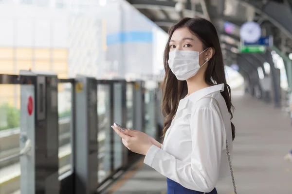 Young Asian professional business woman stands on a sky train station in town wearing a face mask while uses her smartphone to send messages  to the customer office