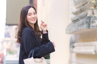 Siyah paltolu güzel, uzun saçlı Asyalı bir kadının portresi, yürüyüş çantası taşıyor, açık havada mutlu bir şekilde gülümsüyor..