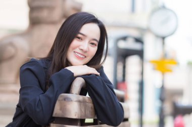 Koyu mavi uzun kollu gömlekli güzel Asyalı iş kadını bir sandalyede oturuyor ve mutlu bir şekilde gülümsüyor ve dışarıdaki sokaktaki kameraya bakıyor..