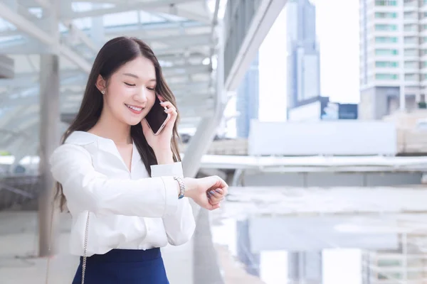 Portrait professional business Asian woman is walking to office or work place while she looks at watch and talks on the phone with smartphone at outdoors in a big city among business buildings as a the background.