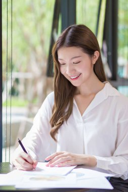 Asyalı kendine güvenen bir kadın çalışmak için beyaz bir not defteri kullanıyor ve masada bir sürü belge var. Evdeki çalışma odasında gülümseyen yüzü..