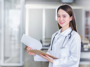 Profesyonel, güzel, genç bir kadın doktor elinde bir dosya, kameraya gülümserken beyaz laboratuvar önlüğü ve hastanede stetoskop takıyor..