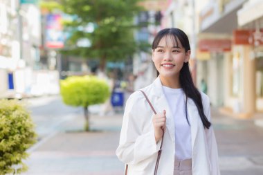 Beyaz gömlek ve omuz çantası giyen kendine güvenen genç Asyalı kadın işe giderken mutlu bir şekilde gülümsüyor..