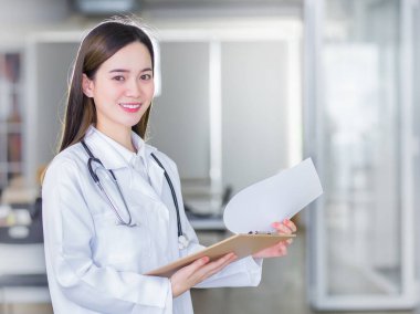 Profesyonel, güzel, genç bir kadın doktor, elinde bir dosya, kameraya gülümserken beyaz laboratuvar önlüğü ve hastanede stetoskop takıyor..