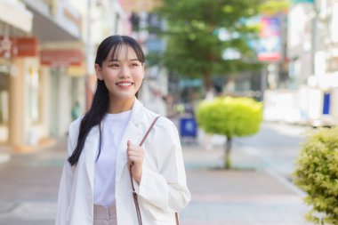 Beyaz gömlek ve omuz çantası giyen kendine güvenen genç Asyalı kadın işe giderken mutlu bir şekilde gülümsüyor..
