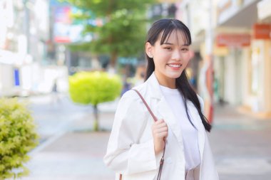 Beyaz gömlek ve omuz çantası giyen Asyalı, kendine güvenen genç iş kadını, işe giderken mutlu bir şekilde gülümsüyor..