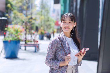 Kahverengi çizgili spor ceket ve omuz çantası takan kendine güvenen genç Asyalı işçi kadın güneşli bir günde işe gidip gelirken mutlu bir şekilde gülümseyen akıllı telefon kullanıyor..
