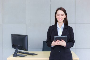 Asian professional working woman in a black suit holds tablet in her hands and confident smiles in office room.