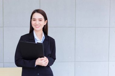 Young Asian professional working woman in a black suit holds clipboard in her hands and confident smiles in office room.
