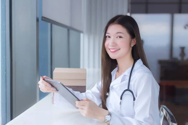 身穿白衣的亚洲女医生坐在医院图书馆的椅子上 在平板电脑上查找一些治疗信息 — 图库照片