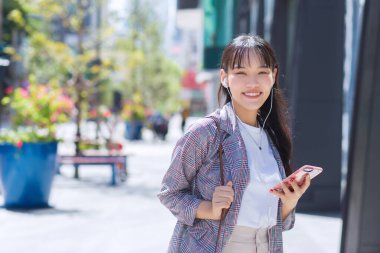 Confident young business Asian working female who wears a brown striped blazer and shoulder bag while using a smartphone with small talk smiles happily as she commute to work through the city on a sunny day.