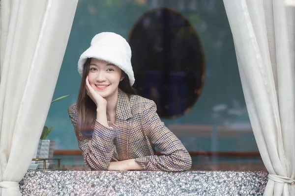 Beautiful Asian female wears a white hat and plaid coat while she sitting near glassed window in New Year and winter theme.