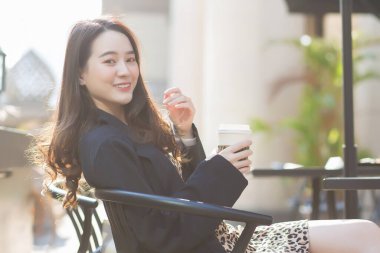 Koyu mavi takım elbiseli Asyalı güzel bir kadın sandalyede oturuyor ve mutlu bir şekilde gülümsüyor. Güneşli bir sabahta kahve dükkanının önünde.