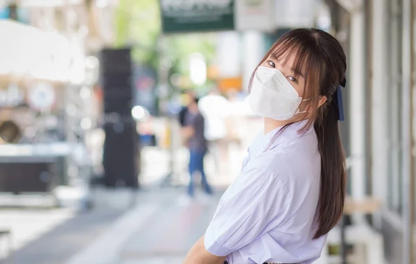 Cute Asian girl student in a uniform wears a protective mask when she is on her way to school through the middle of the city.