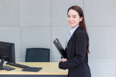 Young Asian professional working woman in a black suit holds clipboard in her hands and confident smiles in office room.