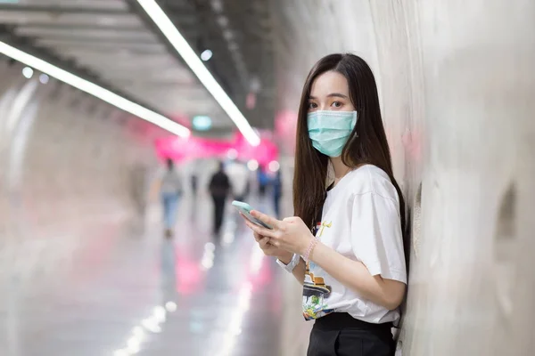 年轻的亚洲女人站在地铁隧道里 手拿智能手机 戴着口罩 这是一种新的正常和健康的生活方式 — 图库照片