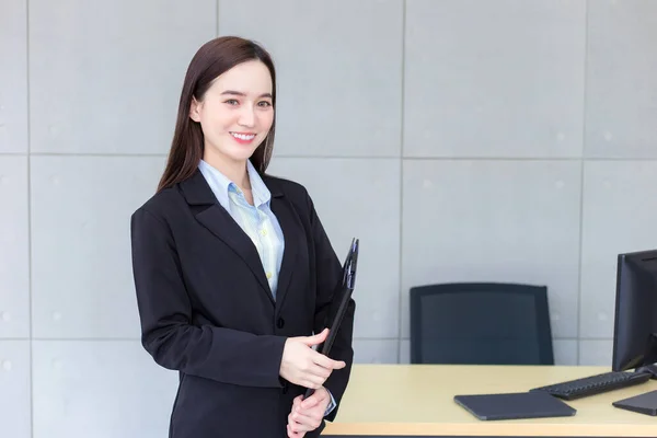 Asian professional working woman in a black suit holds clipboard in her hands and confident smiles in office room.