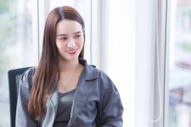 Young business Asian confident working woman who wears dark grey dress sits on chair in office room and works happily.