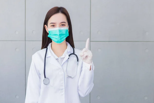 医療面のマスクを身に着けているアジアの女性医師 健康を守るためのゴム手袋は壁に何かを提示することを指しています — ストック写真