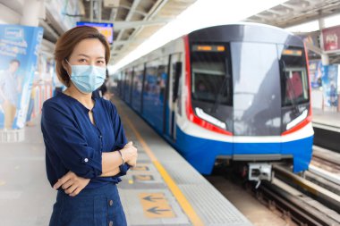 Kısa saçlı ve mavi gömlek giyen Asyalı kadın tren istasyonunda beklerken kol kola giriyor ve sağlık hizmetlerinde COVID-19, kirlilik PM2.5, yeni normal ve koronavirüs koruma yaşam tarzı kavramını korumak için tıbbi maske takıyor..