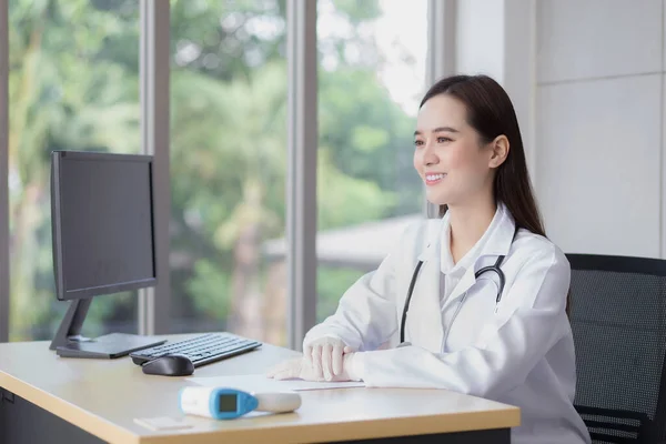 亚洲专业的年轻微笑的女医生坐在医院里翘首以待 桌上有一张纸和一台电脑 — 图库照片