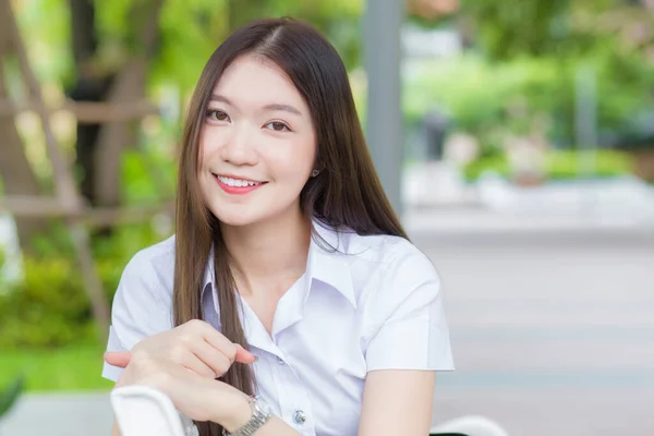 Portrait of an adult Thai student in university student uniform. Asian youg beautiful girl sitting smiling happily at university with a background of garden trees.