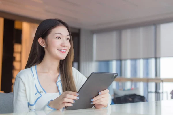 Beautiful young age Asian professional woman with long hair in white shirt holds tablet her smiling happily while she sits on chair in workplace office with glassed building as a background.