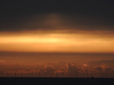 Denizin üzerinde gün doğarken, uzaklarda ve yel değirmenlerinde küçük bir yelkenli görünür.