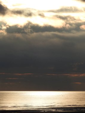 Denizin üzerinde gün batımının görüntüsü, güneş bulutlar tarafından gizleniyor ve suya yansıyor.