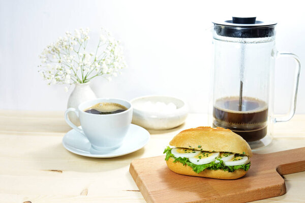 Breakfast with egg slices in a sandwich roll on a wooden kitchen board, coffee maker and cup, light background with copy space, selected focus, narrow depth of field