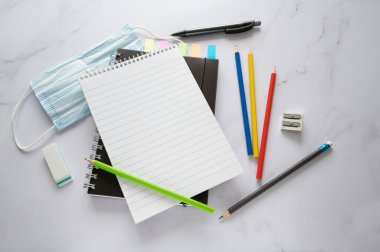 Boş spiral not defteri kopya alanı, pastel boya kalemleri, kovid-19 enfeksiyonuna karşı cerrahi maske ve mermer yüzeyde sabit malzemeler, yukarıdan yüksek açılı görüş, seçilmiş odaklanma