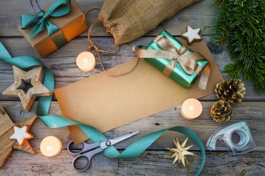 Noel hediyelerini kahverengi kağıt ve turkuaz kurdele, makas, dekorasyon yıldızları ile paketlemek ve gri bir ahşap masa üzerinde mumlar yakmak, ortadaki boşluğu kopyalamak, yukarıdan üst görünüm