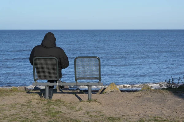 海の上の空の椅子の横に座って後ろから暗い暖かいジャケットの独身男性は 水平線 レクリエーションや愛する人の損失のための喪のための概念 コピースペースに青い水を探している — ストック写真