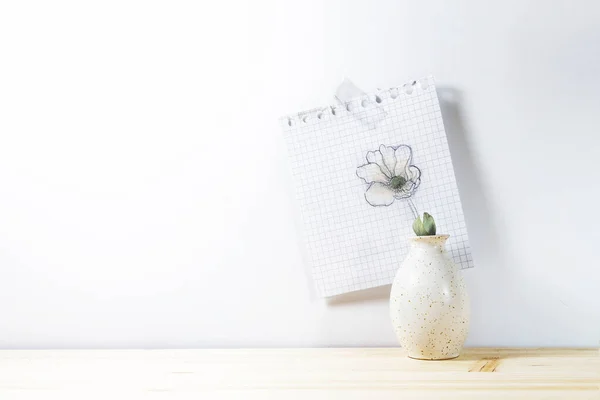 木製のテーブルの上に小さな花瓶とその背後にある紙の上に描かれた花は 白い壁に貼り付けられ 光の背景に寛大なコピースペース — ストック写真