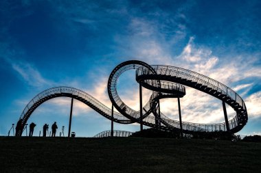 Yürüyebilen roller coaster heykelinde siluet olarak tripodlu fotoğrafçılar. Kaplan ve Kaplumbağa, Almanya 'nın Duisburg şehrinde beyaz bulutlar, sanat kurulumu ve simgesi olan koyu mavi bir gökyüzüne karşı.