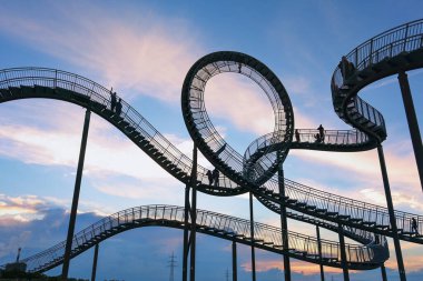 Tiger ve Turtle, yürüyen lunapark treninin merdivenlerindeki siluetler gibi insanlar. Pembe bulutlu mavi gökyüzüne karşı heykel, sanat kurulumu, manzara ve turizm merkezi, Almanya Duisburg,