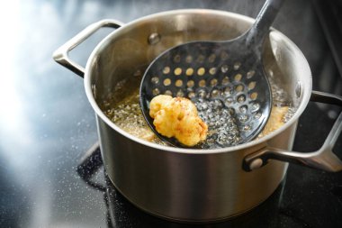 Cauliflower floret on a spatula deep fried in a pot with steaming cooking oil on the stove, creative preparation of a vegetable meal, copy space, selected focus, narrow depth of field