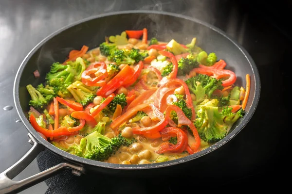 Steaming vegetable curry with chickpeas, broccoli and red bell pepper in coconut milk in a cooking pan on a black stove, healthy meals for a vegan low carb diet, copy space, selected focus, very narrow depth of field