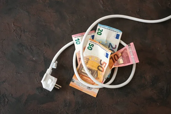 Bundle of euro banknotes tied up with an electric power cable with plug, concept for energy efficiency, power consumption and rising electricity costs, dark background, copy space, selected focus