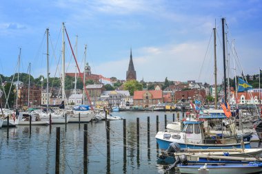 Flensburg, Germany, July 25, 2022:  Old town cityscape with St. Marien church behind the yacht harbor with sailing boats, at the fjord on the Baltic sea, overcast blue sky, copy space, selected focus