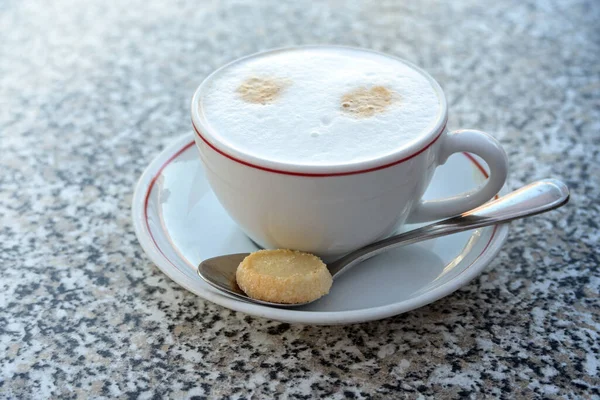 Cappuccino with foamed milk in a white cup and a cookie on a spoon, hot drink served on a granite stone table, copy space, selected focus, narrow depth of field