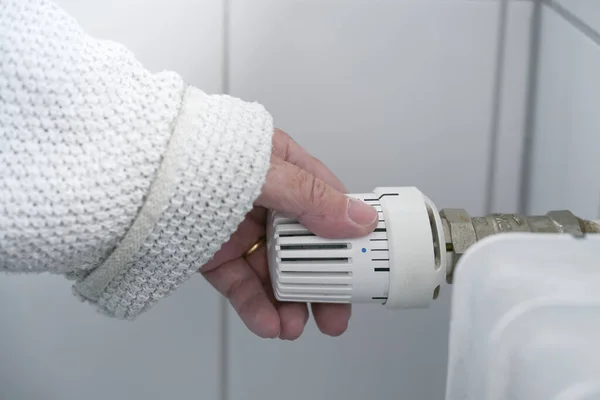 Hand of a woman in warm clothes reduces the heat at the thermostat on the radiator due to the energy crisis with rising prices, copy space, selected focus, narrow depth of field