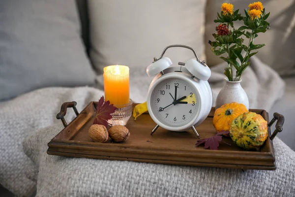 Vintage alarm clock showing one hour fall back after daylight saving time, wooden tray with candle and autumn decoration on a bed with natural blanket and pillows, copy space, selected focus