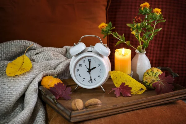 Fall back in autumn after daylight saving time, white alarm clock on a wooden tray with candle and warm colored fall decoration on a red brown couch, copy space, selected focus
